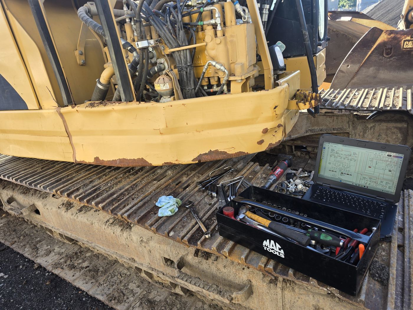 Laptop showing a hydraulic schematic beside a tool tray on the track of an excavator during field diagnostics